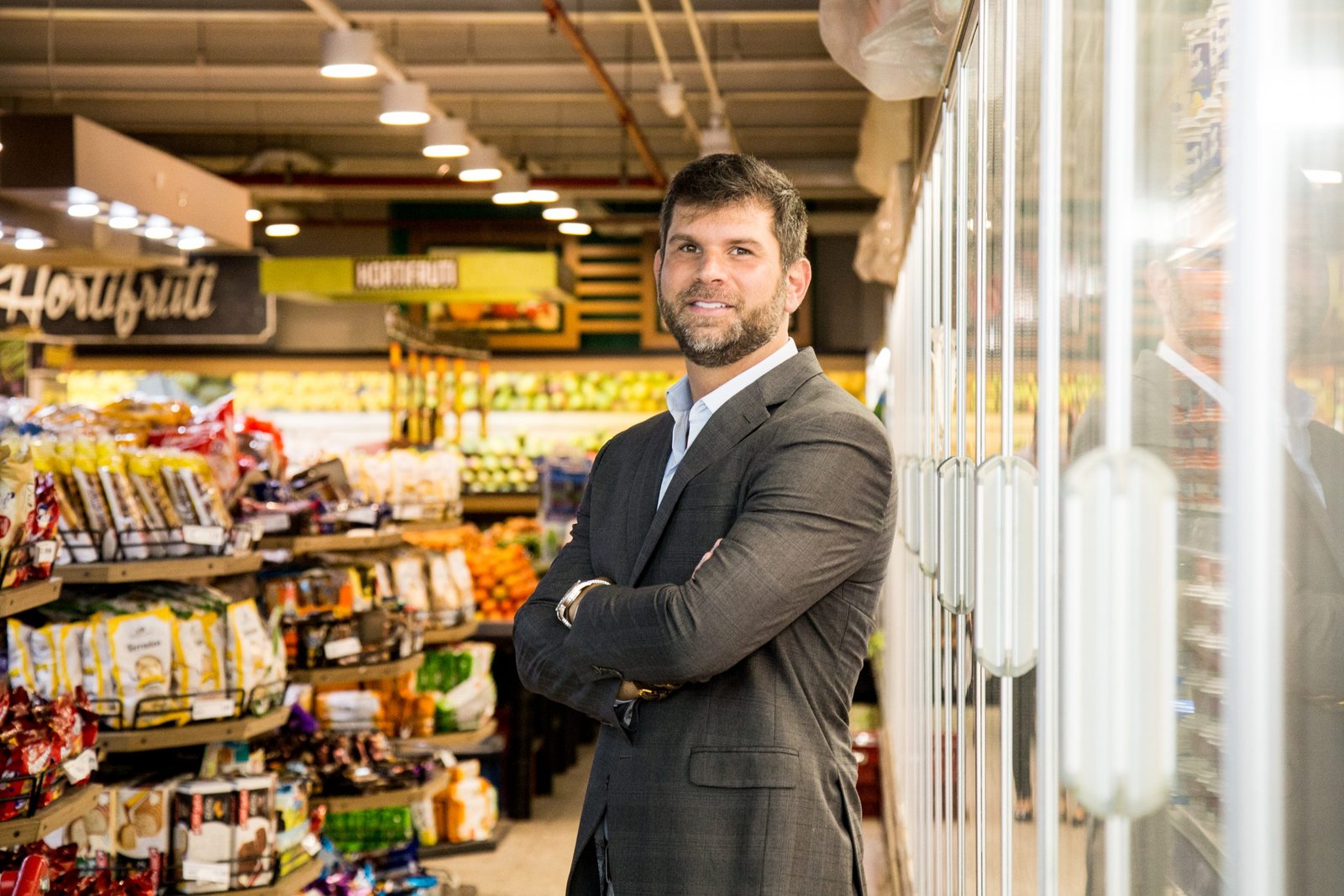 Fábio Queiróz_Homem branco, em pé, de braços cruzados em um corredor de supermercados_Crédito Asserj